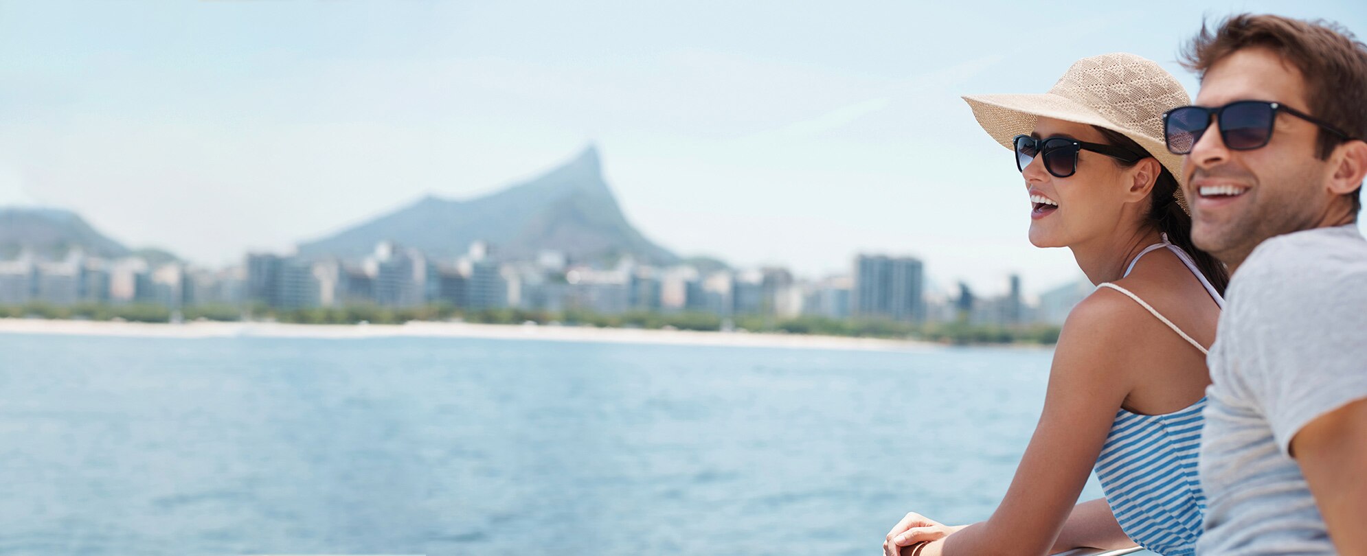 A smiling couple wearing sunglasses on a cruise looking out over the ocean with a city and mountain in the distance.