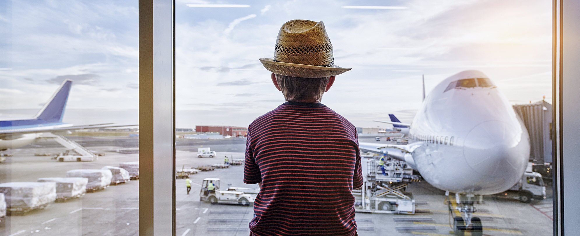 A young boy wearing a hat, looking out of an airport window at a nearby plane. 