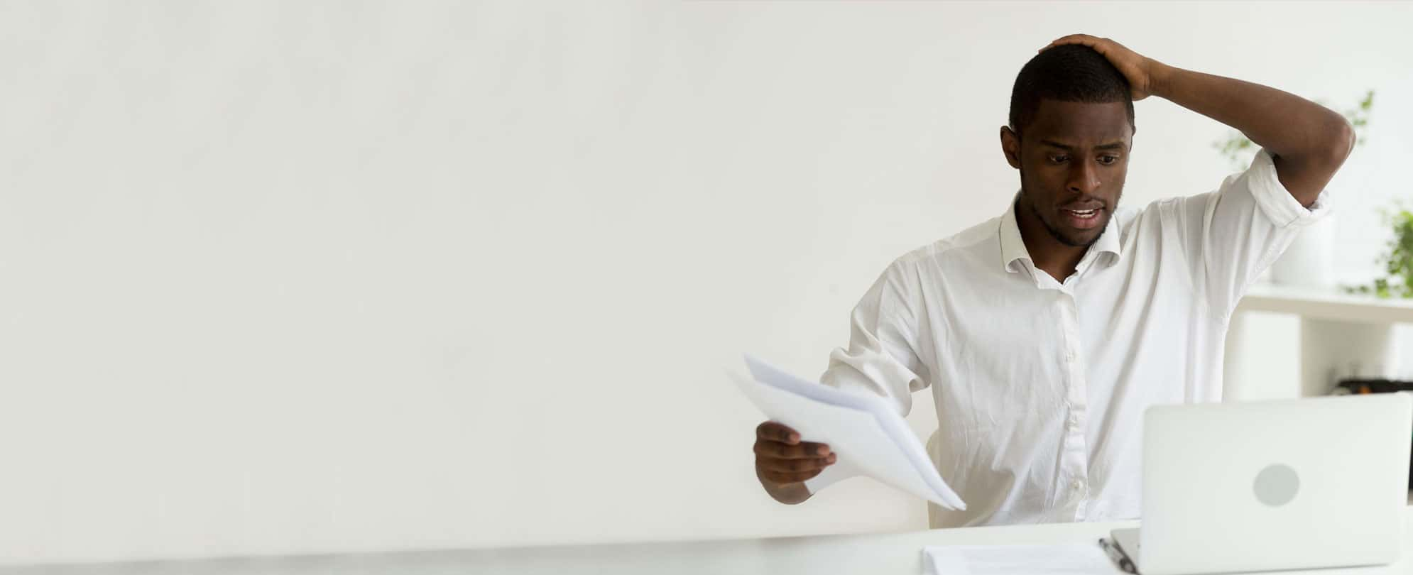 A man looking overwhelmed holding his head in one hand and paperwork in another as he sits at a desk in front of his laptop. 