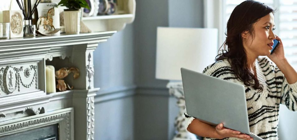 A woman in her living room holding a laptop as she has a conversation on the phone. 