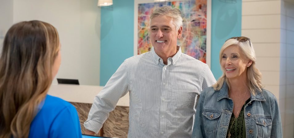 Smiling man and woman stand at a check-in desk, talking to a team member at a Margaritaville Vacation Club resort.