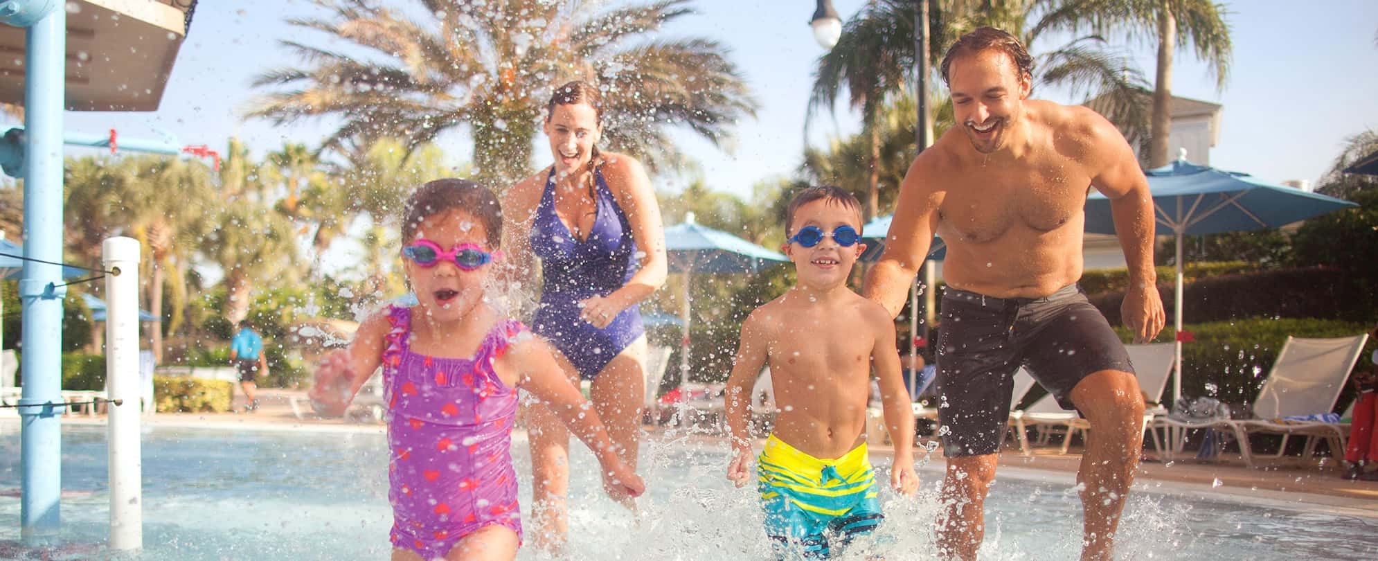 Parents with two young kids wearing goggles run through a splashpad at a timeshare resort.