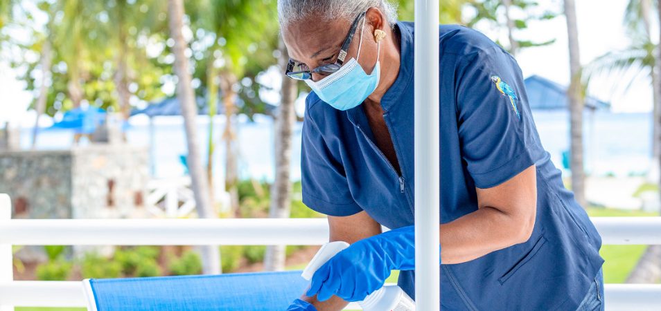 Female employee wearing a blue medical mask and gloves sprays down a table at a Margaritaville Vacation Club resort. 