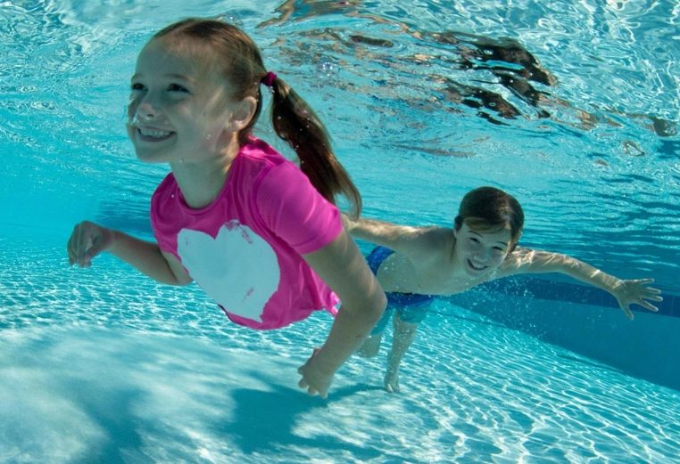 A smiling young girl and boy swim underwater in a pool while enjoying vacation.