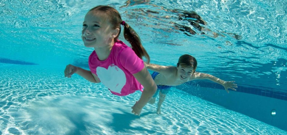 A smiling young girl and boy swim underwater in a pool while enjoying vacation.