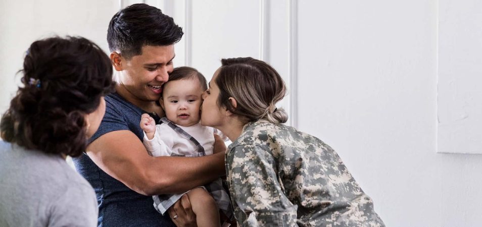 A grandmother lovingly watches a father holding baby while a military mom leans over to kiss the child on the cheek.