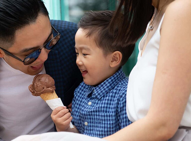 Young boy holding chocolate ice cream cone sits between mom and dad as the dad leans in to take a big bite of ice cream.