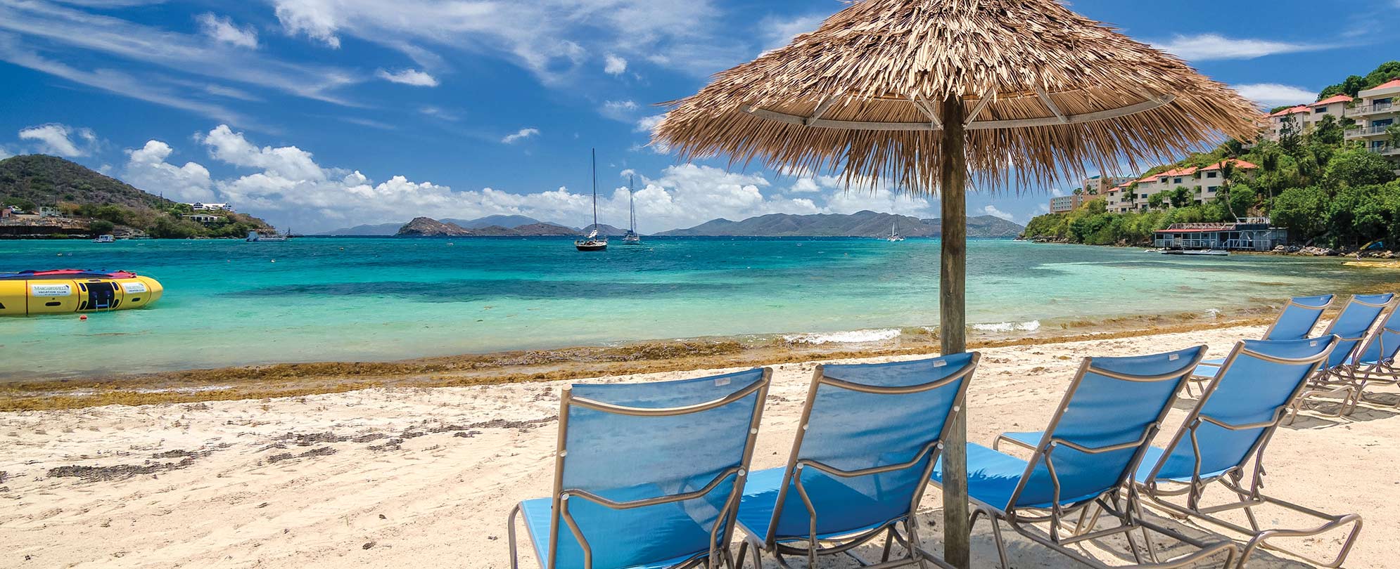 Blue beach chairs sit under a straw umbrella in front of the ocean at Margaritaville Vacation Club by Wyndham - St. Thomas.