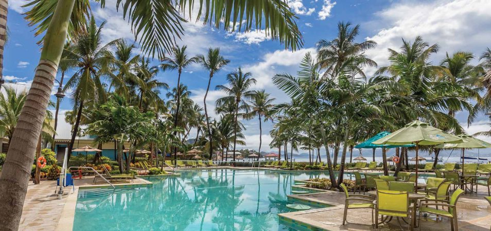 Palm trees and tables with green chairs and umbrellas surround the oceanfront pool at a Margaritaville Vacation Club resort.