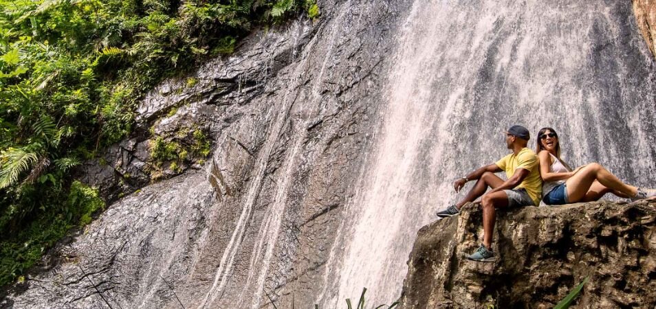 Man and woman sit back-to-back on a rock in front of a waterfall as they explore top things to do in Rio Mar, Puerto Rico.