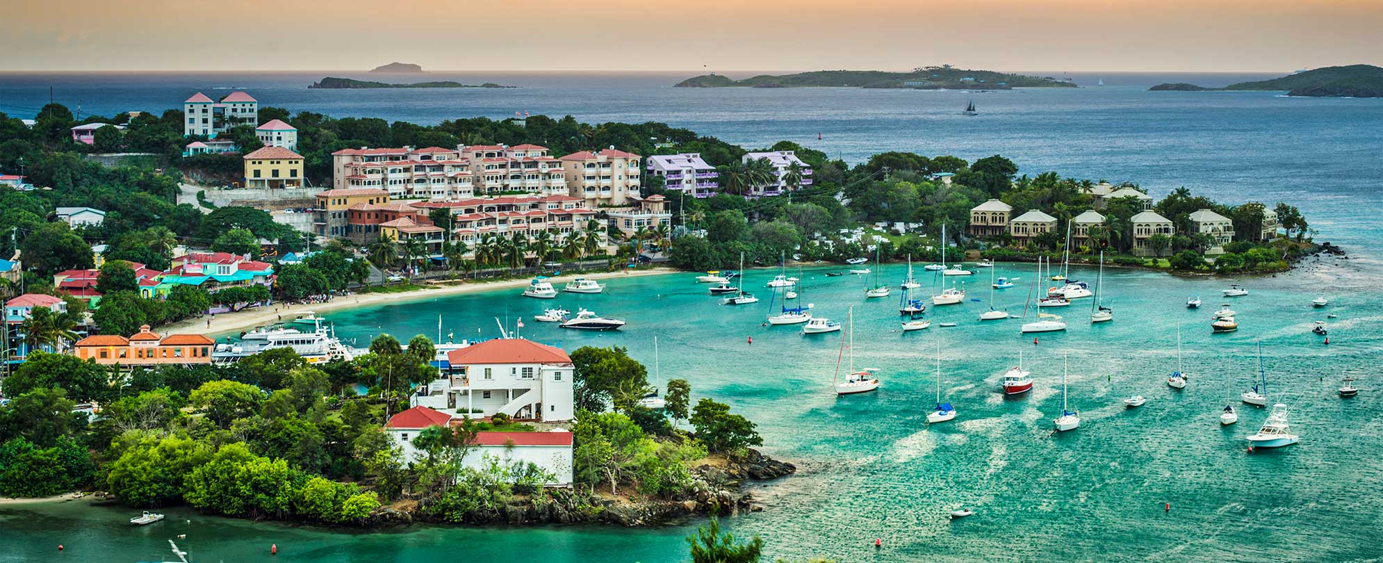 The sun sets over the bright blue ocean surrounding St. Thomas, U.S. Virgin Islands, where sailboats float at an inlet.