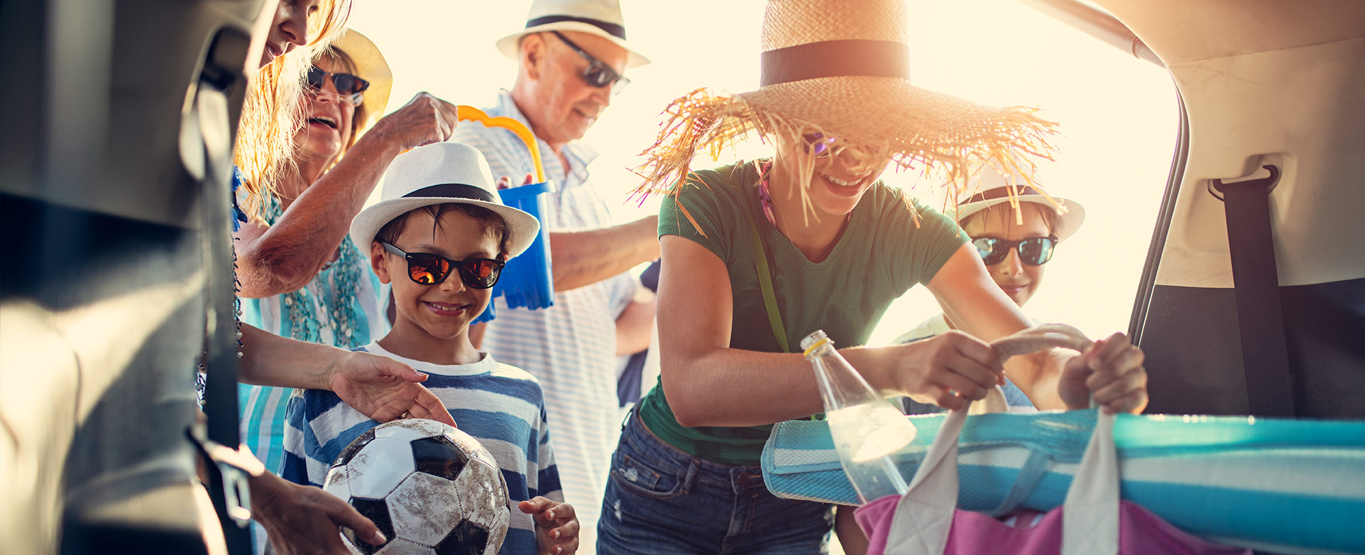 A teenage girl in straw hat and a young boy holding a soccer ball, help their family load up the car for their vacation.